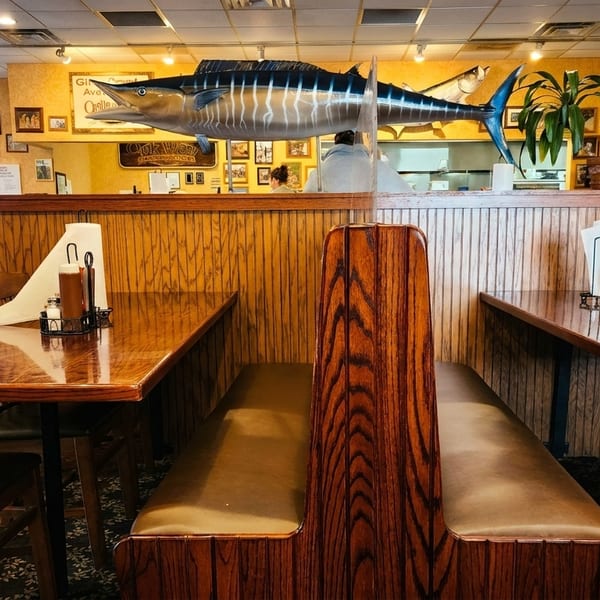 Restaurant interior with wooden booths and tables, a large marlin decoration, and framed pictures on the walls.