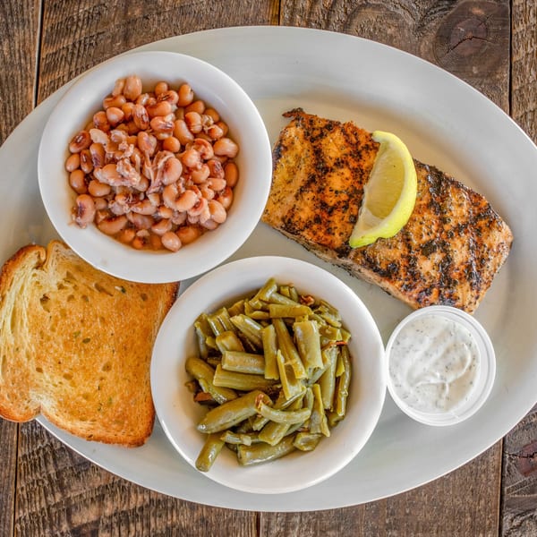 Grilled salmon with a lemon wedge, green beans, pinto beans, and toasted bread on a white plate.