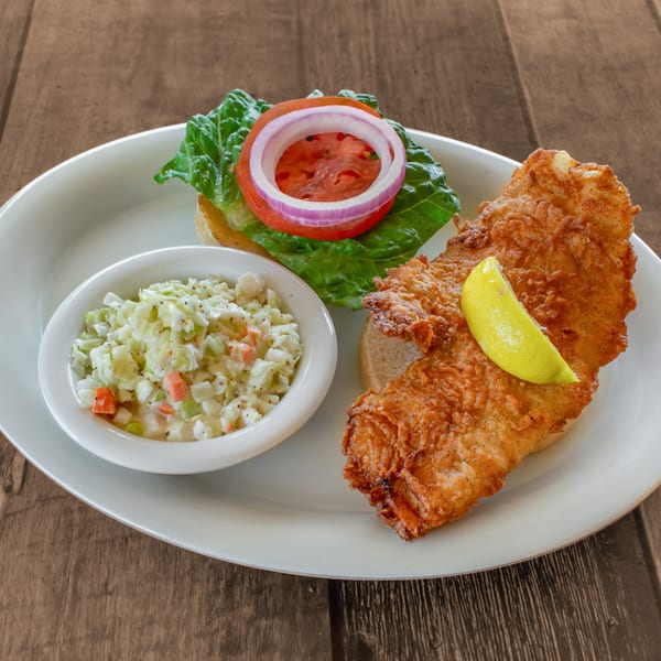 Fried fish sandwich with lettuce, tomato, onion, lemon wedge, and a side of coleslaw.