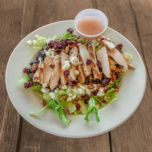 Grilled chicken salad with cranberries, feta, and walnuts, served with dressing on a wooden table.