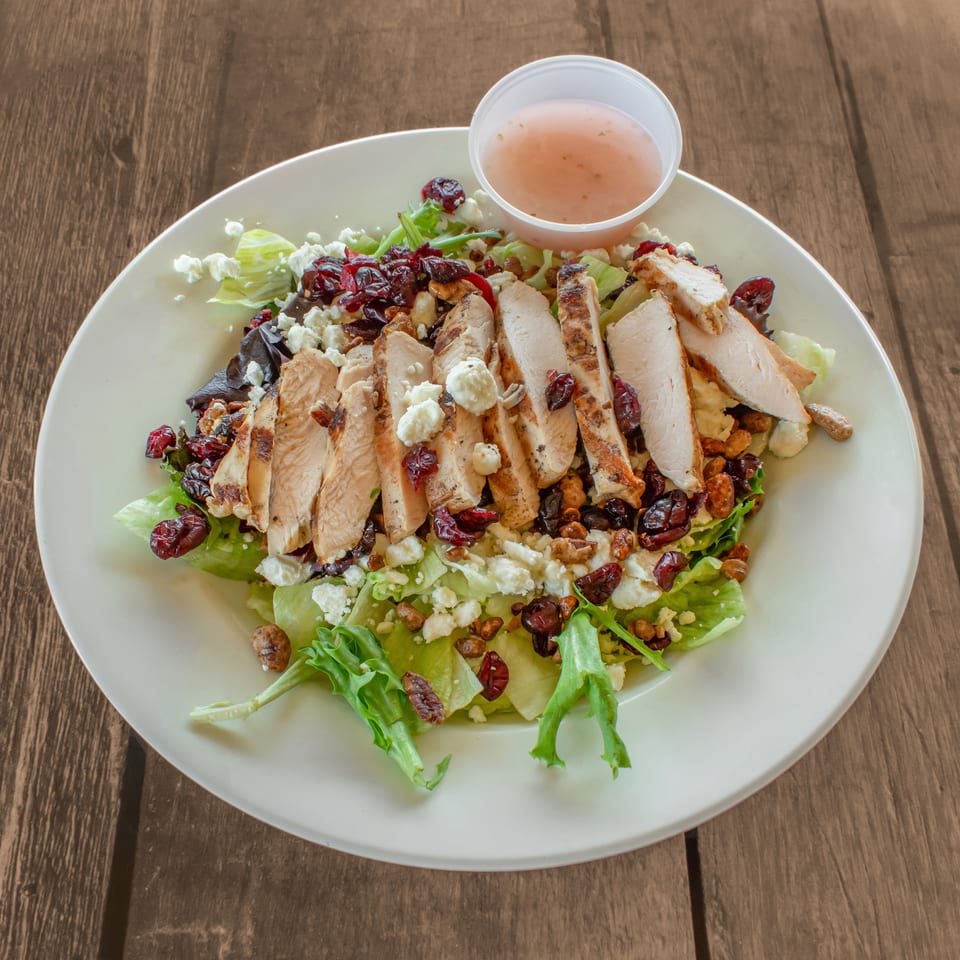 Grilled chicken salad with cranberries, feta, and walnuts, served with dressing on a wooden table.